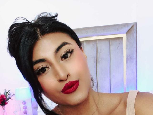 MichelleDenise has dark hair in a messy bun, red lipstick, and long eyelashes. She's wearing a beige top, and her ass is not visible in this shot.