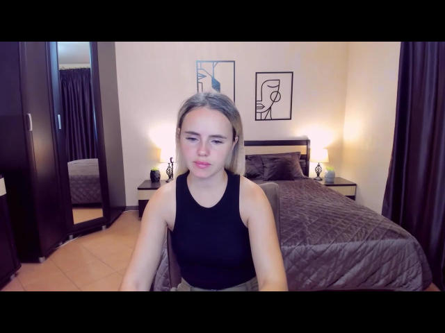 MilanaBloom has blonde hair, small tits, and is wearing a black tank top, sitting in a bedroom with a quilted bed and dark curtains.