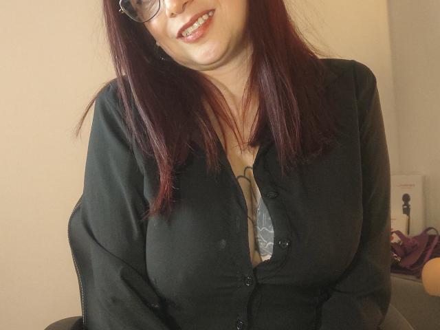 CatalinaMonyque with dark red hair, glasses, and a green blouse, showing her big tits, smiling, and slightly leaning forward. Her blouse is buttoned but revealing.