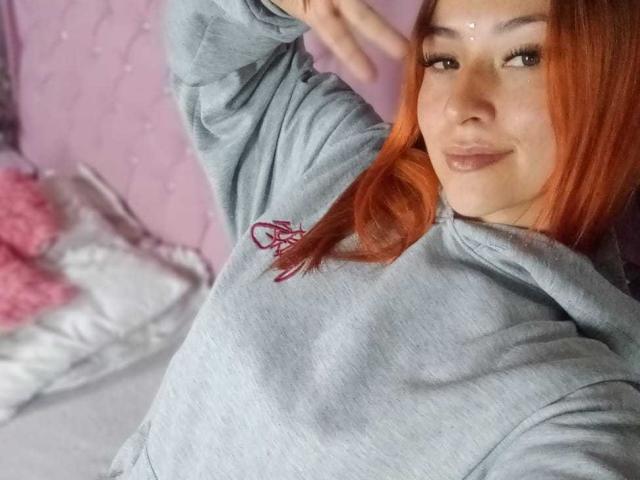 SamanthaCollen with red hair, wearing a gray hoodie, smiling, with a hand up, showing her tits, on a bed with pink and white pillows.
