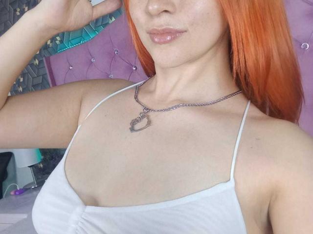 SamanthaCollen with orange hair, wearing a white spaghetti strap top, showing her chest and a silver necklace with a heart-shaped pendant, glossy pink lips, light skin.