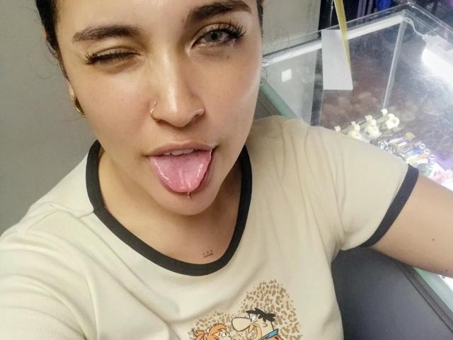AgathaK with dark hair, winking, sticking out her pink tongue, nose ring, and wearing a beige shirt with black trim, in a selfie. Her tongue is slightly drooled.