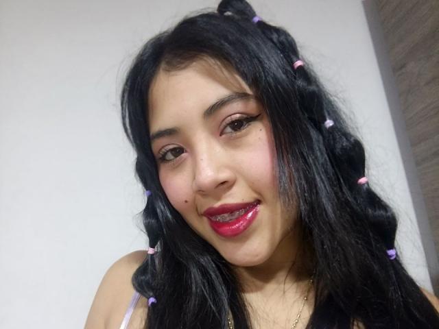 Asiadh has long black hair in braids with pink bows, pink eyeshadow, and pink lipstick, smiling with a small beauty mark on her right cheek. Her tits are small.