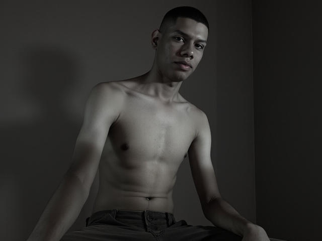 MiloCruz is shirtless with a slim, toned, dark-skinned body, small nipples, and a serious expression. He's wearing dark pants, sitting in dim, shadowy lighting.