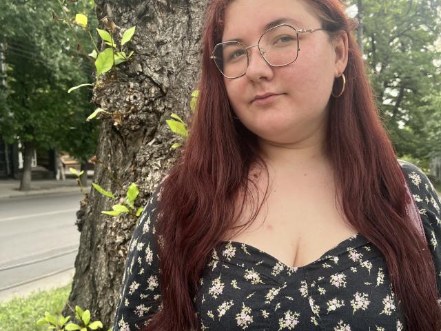 ZlataKissa with red hair, glasses, and hoop earrings, wearing a black floral dress with a cleavage, standing by a tree with green leaves. ZlataKissa's tits are visible.