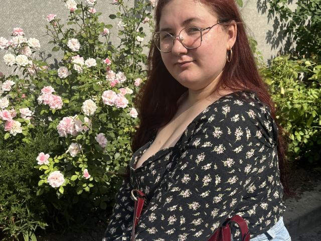 ZlataKissa with red hair, glasses, and hoop earrings, wearing a black floral top with cleavage, standing in front of pink roses. Her tits are partially visible.