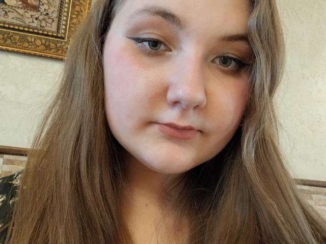 ZlataKissa has long brown hair, light skin, and subtle black eyeliner. Her pink cheeks and closed-lip smile are visible. She's looking straight at the camera.
