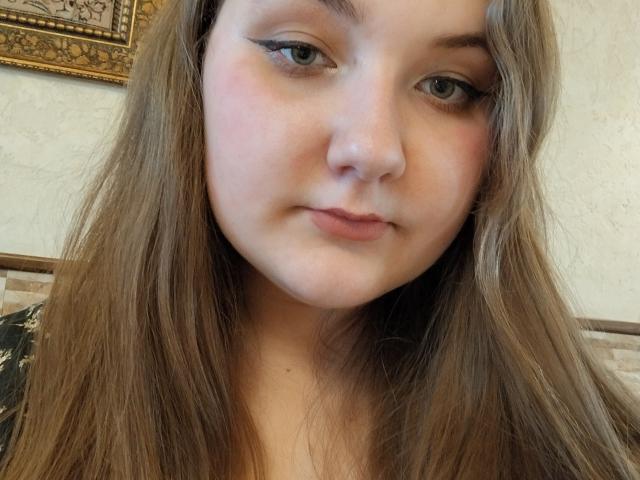 ZlataKissa has long brown hair, light blue eyes, and a slight pink blush on her cheeks. Her lips are closed in a neutral expression. She's wearing a black top.