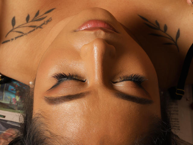 ViickyLee's face, eyes closed, with glossy pink lips, dark eyelashes, and tattoos on her neck. She has tan skin and a slight sheen on her forehead.