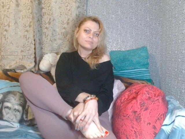 LindaRoutz-hot, blonde, in black off-shoulder top, pink pants, sitting on bed, legs crossed, black bracelet, white socks, red toenails, slight smile, blue pillow, red patterned pillow.
