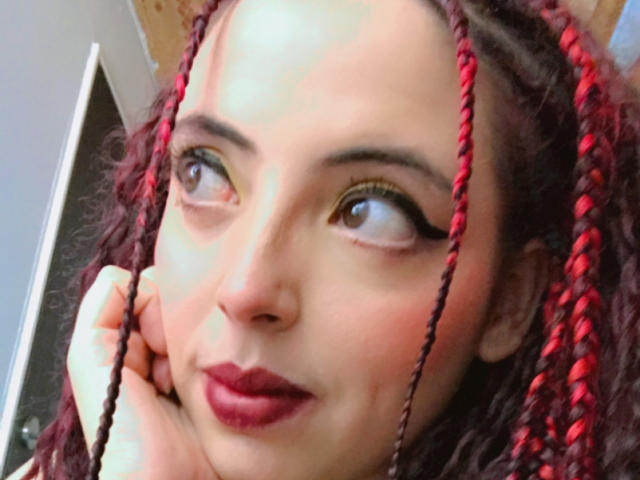 Rosalinnes has red and black braided hair, dark eyeliner, and red lipstick. She looks to the side with a slight smile, touching her cheek. Her skin is fair.