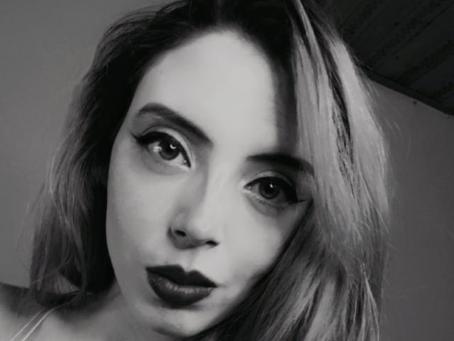 Rosalines has dark, thick eyeliner, black lipstick, and a slight pout. Her gray, wavy hair falls over her face. Her nose is pierced. Black-and-white photo.
