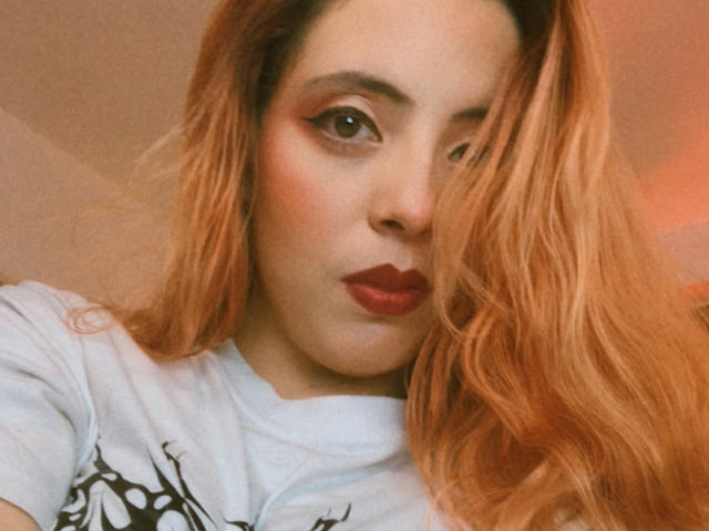 Rosalinnnes with long, wavy, orange hair, full red lips, and smoky eye makeup, wearing a white t-shirt, looking at the camera with a slightly pouty expression.