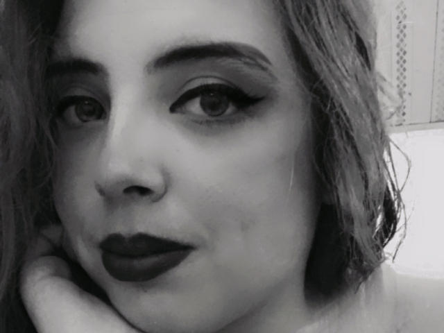 Rosalinnes has dark, winged eyeliner, thick eyebrows, and dark lipstick. Her head is slightly tilted with a hand near her face in a black-and-white selfie.