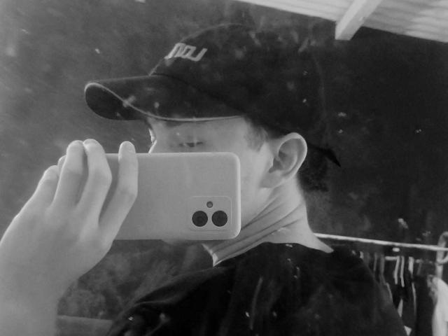JonathanMoreau with a baseball cap taking a selfie in a mirror, holding a white iPhone, reflection shows his face and hand, wearing a dark shirt, black background.