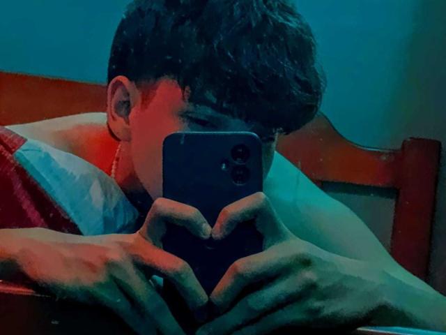 JonathanMoreau with curly brown hair, holding a phone, making a heart shape with his hands, sitting in bed, blue and red light. His phone has a triple-camera setup.