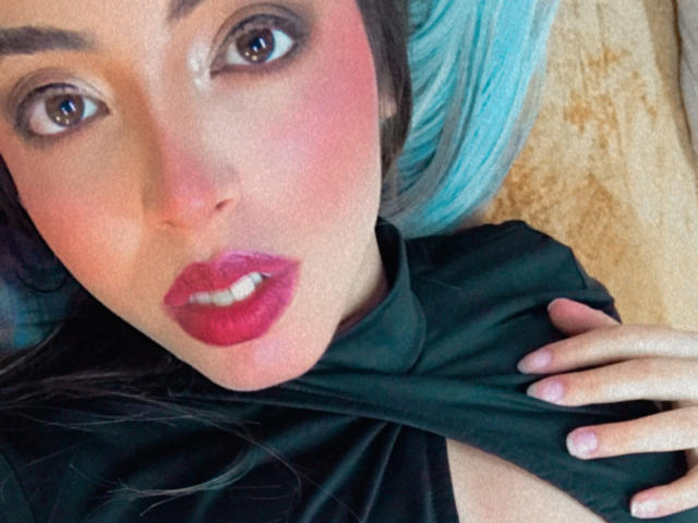 Rosalinnes has pink lipstick, red blush, and blue hair, lying down with black shirt pulled up, exposing her tits, brown eyes looking at camera, hand on shirt.