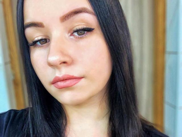 MargoRoiss tem cabelo preto liso, olhos castanhos com delineador preto grosso e batom rosa-avermelhado. Sua pele é clara com um leve brilho, e ela está vestindo uma blusa preta..
