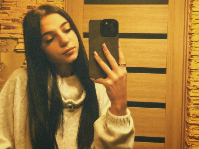 OliviaFoxy with long black hair, wearing a grey sweater, holding a phone in a mirror selfie, her ass is not visible, her eyes are down, she's looking cute.