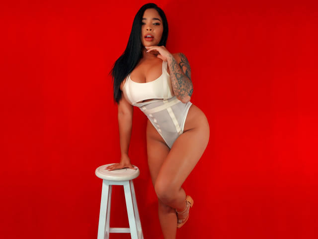 VickyGreys with black hair, tan skin, tattoos, and large tits, wearing a white high-cut thong, leaning on a stool against a red background, with her finger near her mouth.