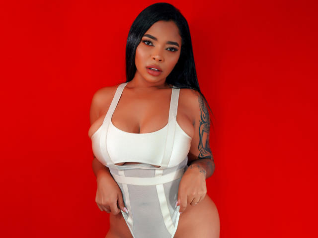 VickyGreys with long black hair, medium brown skin, tattoo on right arm, wearing white cut-out one-piece, showing large tits and curvy ass, against bright red background.