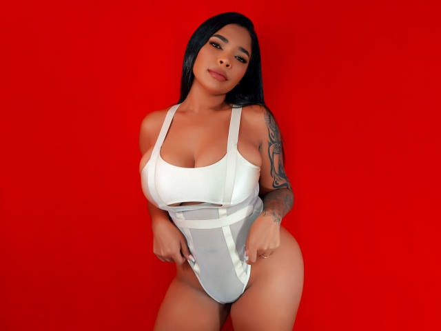 VickyGreys with black hair, tan skin, and tattoos, wearing a white one-piece, showing her big tits and curvy ass against a red background. She's looking at the camera.