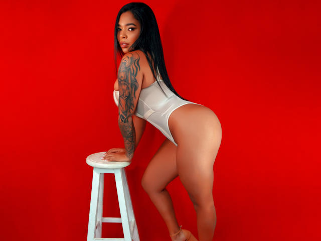 VickyGreys with long black hair, tattoos on her right arm, white one-piece, leaning on white stool, red background, looking over shoulder, showing her ass and butt.