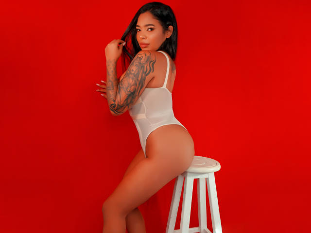 VickyGreys with tattoos, wearing a white thong bodysuit, sitting on a white stool, against a red background, showing her ass and looking over her shoulder.