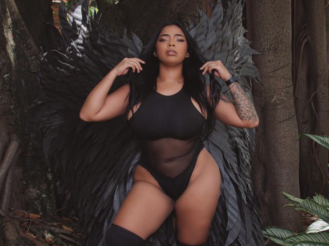 VickyGreys with black wings, wearing a sheer black leotie, showing her big ass and big tits, long black hair, tattooed arm, and black thigh-highs, standing in a forest.