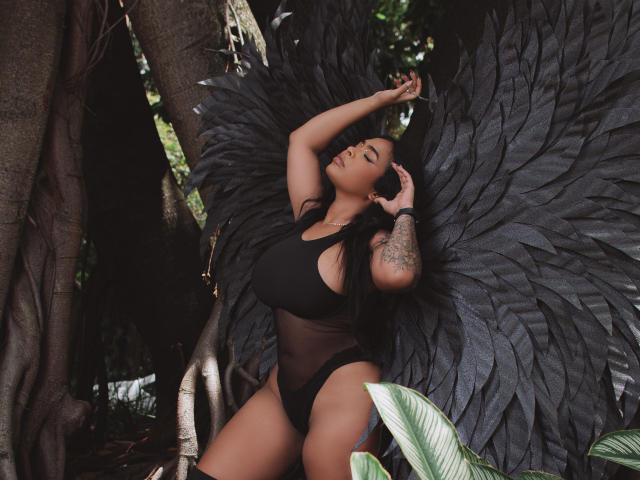 VickyGreys with black wings, wearing a sheer black bodysuit, showing big boobs and a tattooed arm, leaning against a tree, looking sultry. Her black hair is flowing.