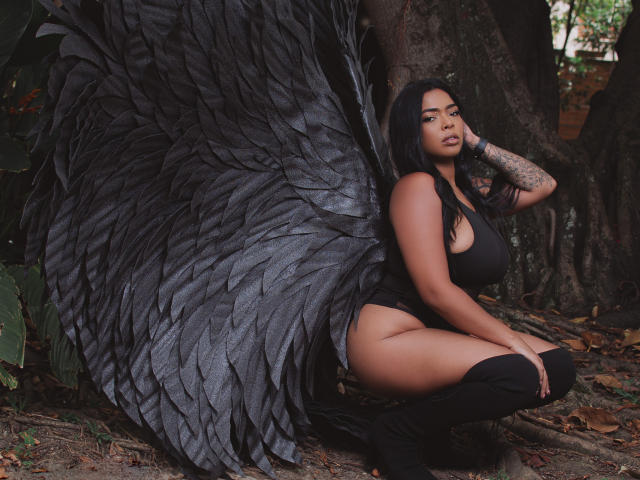 VickyGreys with large black wings, squatting, wearing a black tank top and thigh-high boots, showing her big ass and tattoos, in a forest. Her dark hair is loose.