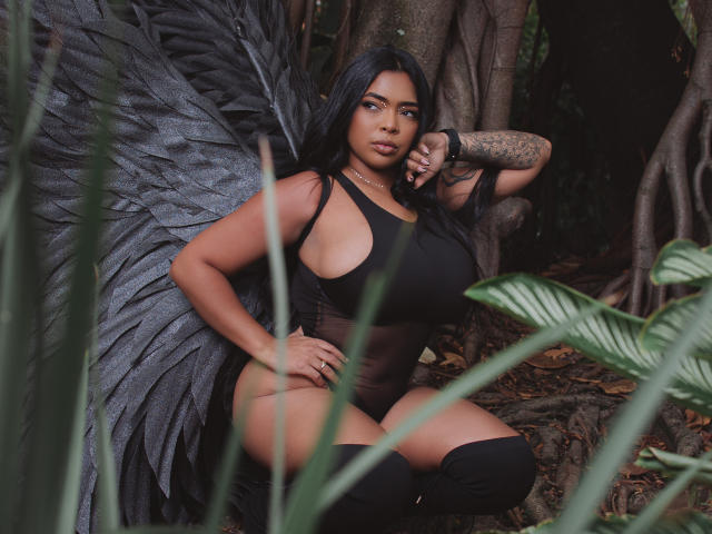 VickyGreys with big tits, black wings, and a tattooed arm, crouching in a forest, wearing a black tank top and black thigh-highs, looking confident.