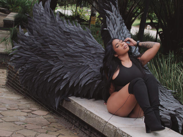 VickyGreys with black wings, sitting on a stone ledge, wearing a black tank top, black thigh-high boots, long black hair, tattooed arm, and a relaxed, sultry expression.