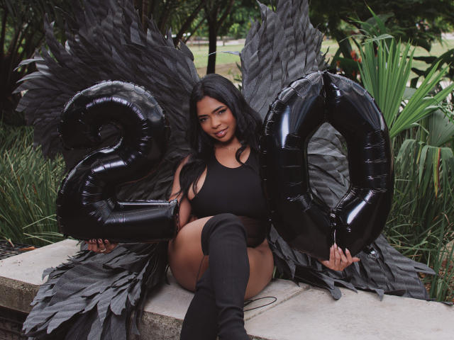 VickyGreys with black wings, black latex number 20 balloons, black tank top, and black thigh-high boots, sitting on a stone, smiling, long black hair, brown skin, outdoors.