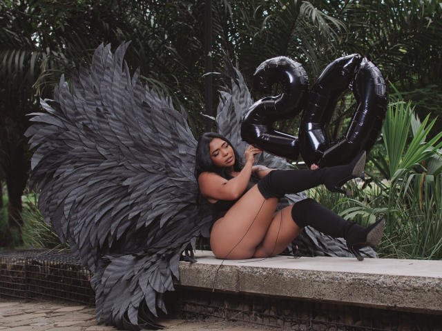 VickyGreys with huge black feather wings, black latex boots, and holding black balloon numbers, sitting by a tiled pool, her bare ass and pussy visible. Her long black hair.