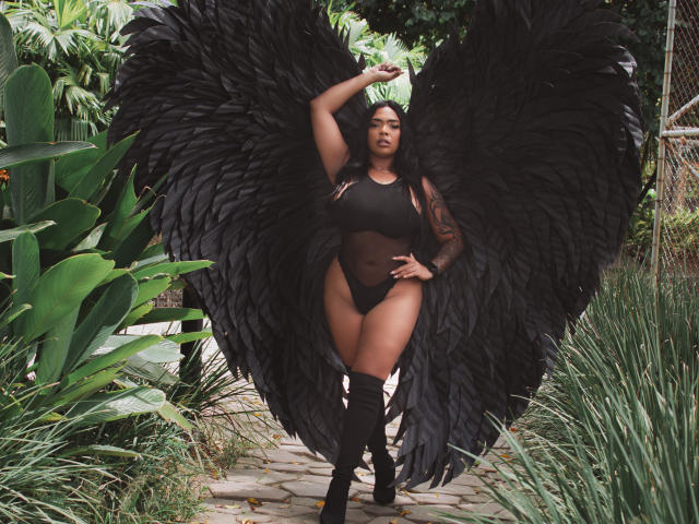 VickyGreys with huge black wings, black mesh bodysuit, black boots, dark skin, tattooed arm, standing on stone path, green foliage around, hand on head, confident expression.