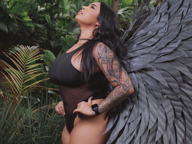 VickyGreys with big tits, black sheer one-piece, tattooed arm, black wristband, gray feather wings, long black hair, standing in jungle, looking up, hands on hips.