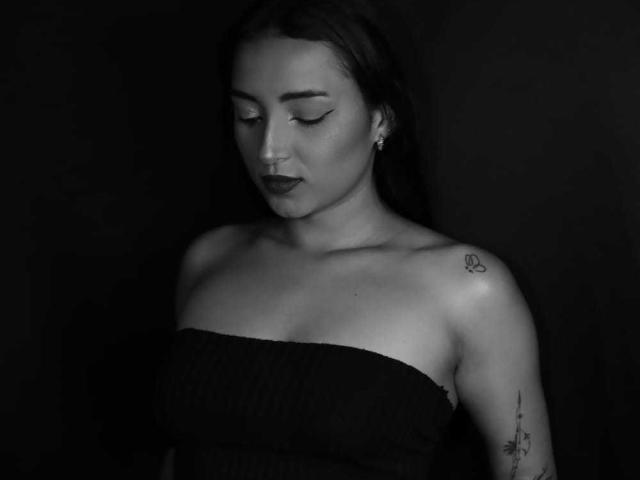 Black and white photo of MaraLewis with eyes closed, wearing a strapless dark top, showing tattoos on her shoulder and upper arm, and a nose ring.