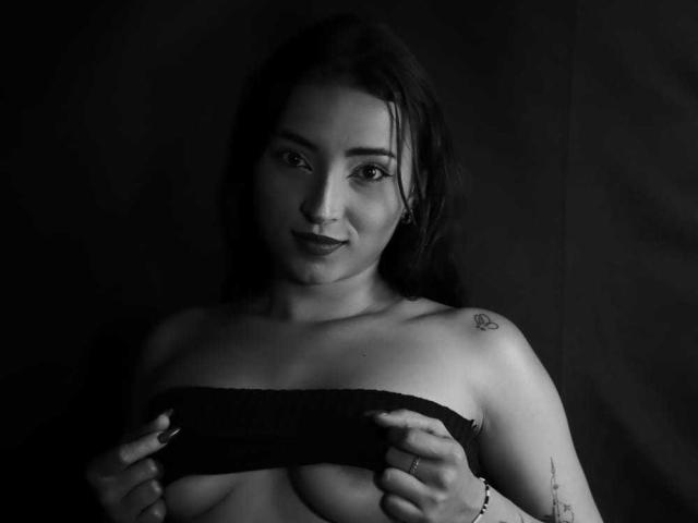 Black-and-white photo of MaraLewis with dark hair, holding her bare tits with a slight smile, tattoo on right shoulder, and a ring on her left hand.