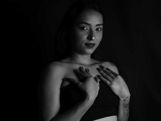 MaraLewis with dark nails, hands on chest, strapless top, tattoos, subtle smile, black-and-white, dark background, looking at camera. Boobs partially covered, dark lipstick, bracelet on right wrist.