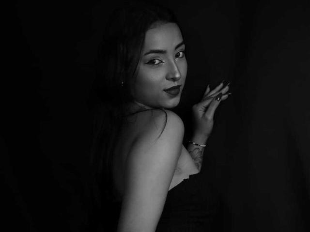 MaraLewis with dark lipstick, long hair, and a bracelet, looking over her shoulder, showing her bare back and tits in a black-and-white photo.