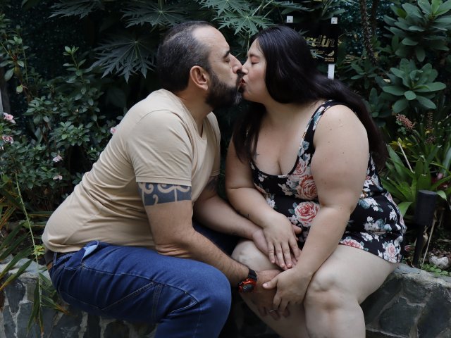 SarahMat, una donna formosa con un seno prosperoso, vestita con un abito floreale, bacia un uomo barbuto con un tatuaggio tribale sul braccio, seduto su una panchina di pietra in un giardino..