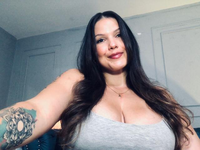 SimoneCallen with long dark hair, gray tank top, and large boobs, smiling at the camera, tattoo on her right arm, small cross necklace, light skin, pink lipstick.
