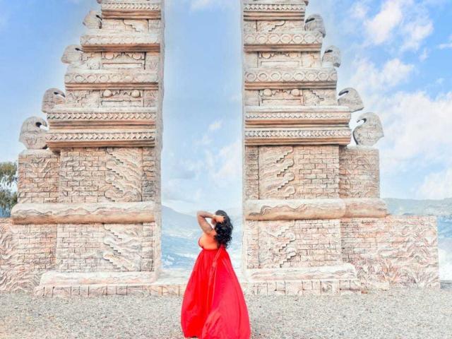 VickyGreys with red dress, back to camera, standing in front of tall, ornate stone towers, hands in curly hair, blue sky with clouds. Gravel ground. Rock carvings.