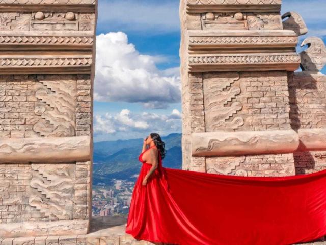 VickyGreys in a red dress, holding a red fabric against ancient stone pillars with detailed carvings, looking over a mountainous cityscape under a blue, cloudy sky.