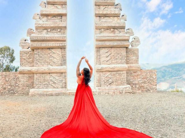 VickyGreys in a red dress, back to camera, arms raised, standing in front of ornate stone gate, dress pooling on gravel, blue sky, white clouds, and distant hills.