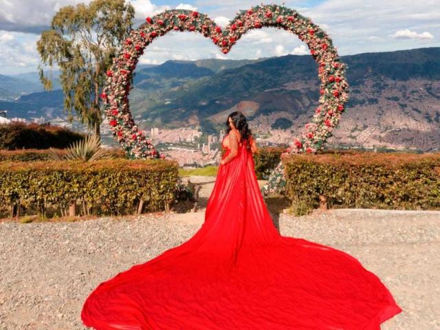 VickyGreys in a red, floor-length, sheer dress with a massive, flowing train, standing before a heart-shaped flower arrangement, back to the camera, in a sunny, mountainous setting.
