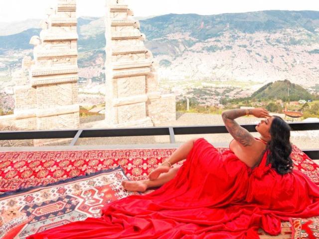 VickyGreys with tattoos, red sari, lying on patterned rugs, hands over head, looking at tall stone towers, mountain city in background, sunlight, relaxed, sensual, elegant.