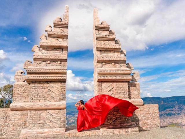 VickyGreys with a red cloth flying, standing between two tall, stone, ornate, ancient temple structures, under a bright blue sky with white clouds.