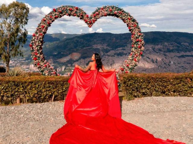 VickyGreys with red dress and long red skirt, holding fabric, standing in front of heart-shaped flower arrangement, mountains in background, clear sky, gravel path, lush greenery.